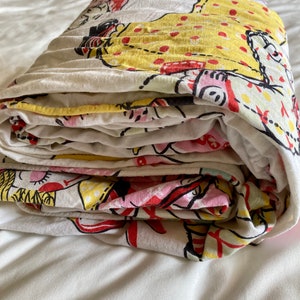 May include: A folded white cotton sheet with a colorful print of cartoon figures in red, yellow, and pink. The figures are wearing dresses and have polka dots.