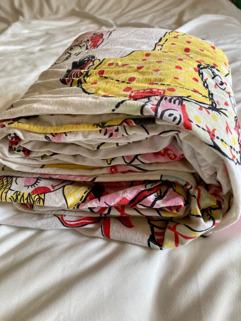 May include: A folded white cotton sheet with a colorful print of cartoon figures in red, yellow, and pink. The figures are wearing dresses and have polka dots.