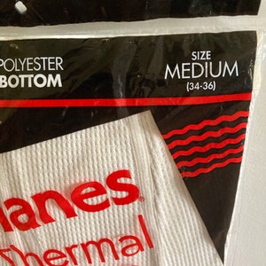 Peut inclure: Un sous-v&ecirc;tement thermique blanc avec le texte "Hanes Thermal Bottom" en rouge et une &eacute;tiquette de taille indiquant "Taille M (34-36)".