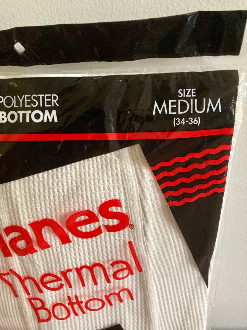 Peut inclure: Un sous-v&ecirc;tement thermique blanc avec le texte "Hanes Thermal Bottom" en rouge et une &eacute;tiquette de taille indiquant "Taille M (34-36)".