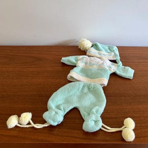 May include: A baby's knitted outfit in shades of mint green, white, and yellow, including a sweater, pants, and a hat. The sweater has horizontal stripes, and the hat and pants have pom-pom accents. The outfit is displayed on a wooden surface.