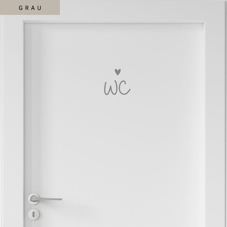 Op de afbeelding: Een witte deur met een grijze sticker met de tekst WC in een handgeschreven lettertype, bekroond met een klein grijs hartje. De deur is voorzien van een zilveren metalen deurklink en een bijpassend sleutelgatplaatje aan de linkeronderkant.