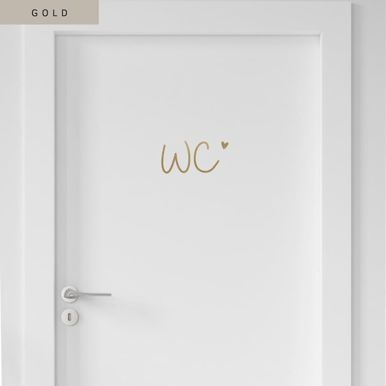 Op de afbeelding: Een witte deur met een goudkleurige sticker met de letters WC in een handgeschreven lettertype, vergezeld van een klein gouden hartje. De deur is voorzien van een zilverkleurige metalen deurklink en een rond sleutelgatplaatje aan de linkerzijde.