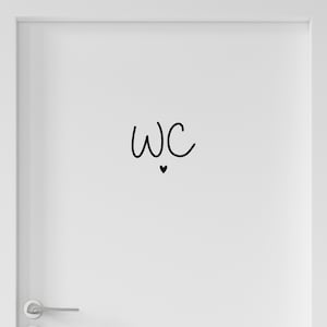 Op de afbeelding: Een witte deur met een zwarte vinylsticker met de letters WC in een handgeschreven lettertype, direct boven een klein, effen zwart hartsymbool geplaatst. De deur is voorzien van een zilverkleurige metalen deurklink in de linkerbenedenhoek.