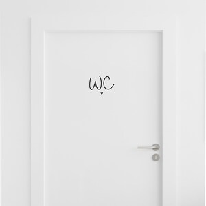 Op de afbeelding: Een witte deur met een zwarte vinylsticker met de letters WC in een handgeschreven lettertype, geplaatst boven een klein, effen zwart hartje. De deur is voorzien van een zilverkleurige horizontale deurklink en een bijpassend sleutelgat.