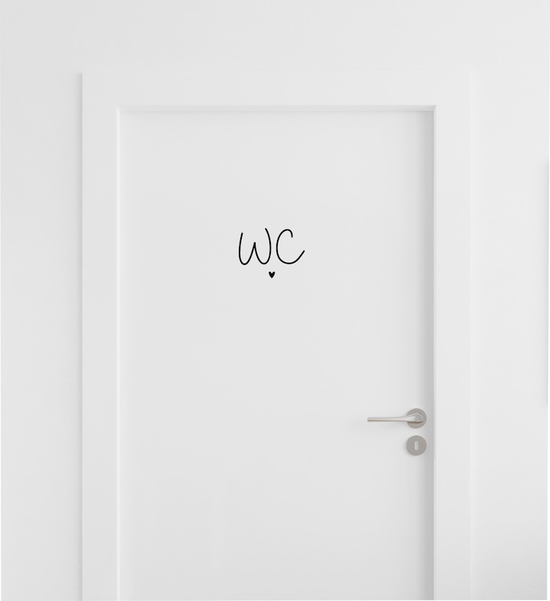 Op de afbeelding: Een witte deur met een zwarte vinylsticker met de letters WC in een handgeschreven lettertype, geplaatst boven een klein, effen zwart hartje. De deur is voorzien van een zilverkleurige horizontale deurklink en een bijpassend sleutelgat.
