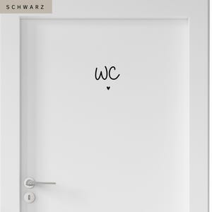 Op de afbeelding: Een witte deur met een zwarte vinylsticker met de letters WC in een handgeschreven lettertype, geplaatst boven een klein zwart hartsymbool. De deur is voorzien van een zilverkleurige metalen deurklink en een bijpassend rond sleutelgatplaatje.