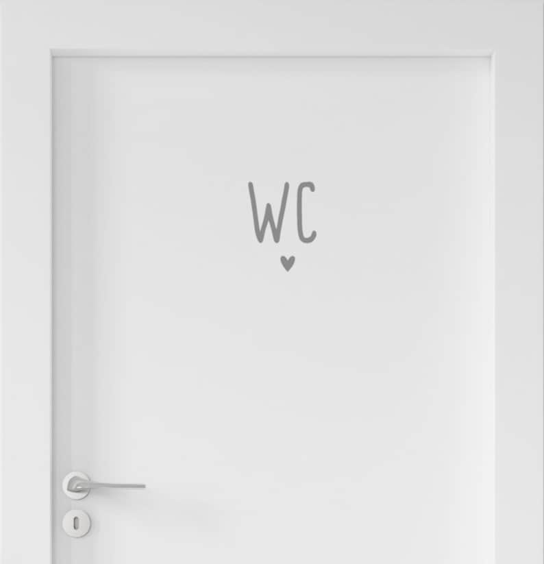 Peut inclure: Une porte blanche dot&eacute;e d'une poign&eacute;e en m&eacute;tal argent&eacute; et d'un autocollant gris au centre. L'autocollant affiche les lettres WC dans une police simple et arrondie, plac&eacute;es juste au-dessus d'une petite ic&ocirc;ne de c&oelig;ur gris solide centr&eacute;e.