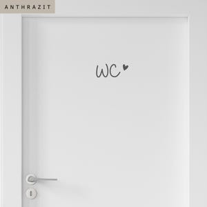 Op de afbeelding: Een witte deur met een donkergrijze sticker met de tekst WC en een klein hartsymbool. De deur is voorzien van een zilverkleurige metalen klink en een bijpassend rond sleutelgatplaatje. Het woord ANTHRAZIT staat in een vakje linksboven.