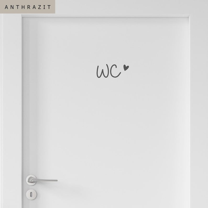 Op de afbeelding: Een witte deur met een donkergrijze sticker met de tekst WC en een klein hartsymbool. De deur is voorzien van een zilverkleurige metalen klink en een bijpassend rond sleutelgatplaatje. Het woord ANTHRAZIT staat in een vakje linksboven.