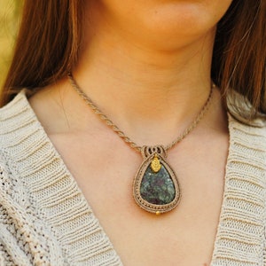 May include: A macramé necklace with a teardrop-shaped pendant and a dark green stone. The pendant hangs from a braided cord. The necklace is worn over a beige knitted sweater.