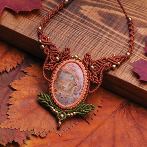 May include: A brown macrame necklace with a large oval-shaped agate stone pendant. The stone has a pink, white, and brown pattern. The necklace is lying on a bed of brown and red autumn leaves.