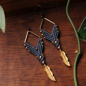 May include: A pair of dangle earrings featuring dark gray macrame cord knotted in a chevron pattern around a diamond-shaped brass frame. Each earring is accented with small brass beads and finished with a dangling brass feather charm at the base.