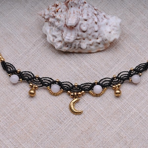 Peut inclure: Un collier en macramé noir avec des accents dorés et un pendentif en forme de croissant de lune. Le collier comporte de petites perles roses pâles rondes et des breloques dorées en forme de goutte. Un coquillage est en arrière-plan.