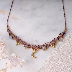 May include: A brown macrame necklace featuring a central gold-toned crescent moon charm, interspersed with small metallic beads and four round, translucent grey stones. The cord is braided and thin, creating a delicate, symmetrical pattern.