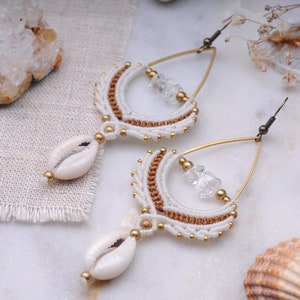 May include: A pair of white and brown macrame earrings with gold accents. The earrings feature a seashell charm and a clear quartz crystal.