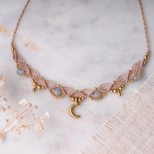 May include: A macrame necklace featuring a light beige cord woven into a scalloped pattern, accented with small gold-toned beads and round, pale blue translucent stones. A gold-toned crescent moon charm hangs from the center of the necklace.