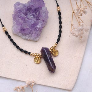 May include: A purple amethyst crystal pendant necklace with gold accents on a black cord. The necklace is displayed on a white fabric surface.