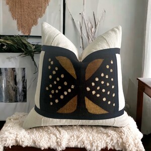 May include: A white linen pillow with a black and gold geometric pattern. The pattern features a stylized butterfly design with gold dots on a black background.