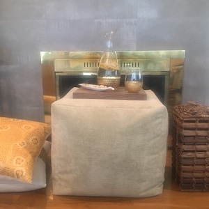 May include: A beige ottoman with a wooden top, a glass pitcher with a gold lid filled with water and lemon slices, a gold glass, and a white ceramic dish with small white stones on top. The ottoman is in front of a fireplace with a brass surround.