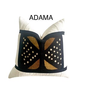 May include: A white pillow with a black and brown geometric pattern. The pattern features a stylized design with gold accents and white dots. The pillow has a black border and the text "ADAMA" is printed above the pillow.