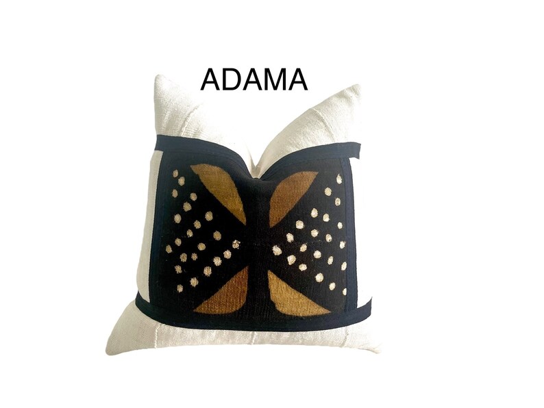May include: A white pillow with a black and brown geometric pattern. The pattern features a stylized design with gold accents and white dots. The pillow has a black border and the text "ADAMA" is printed above the pillow.