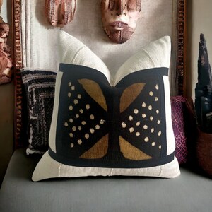 May include: A white throw pillow with a black and gold geometric pattern. The pattern features a large, stylized X shape with gold accents and black dots.