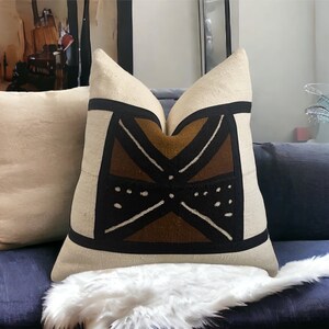 May include: A decorative throw pillow with a geometric pattern in brown, black, and white on a beige background. The pillow is sitting on a blue couch with a white faux fur throw.