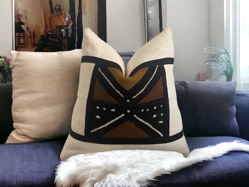 May include: A decorative throw pillow with a geometric pattern in brown, black, and white on a beige background. The pillow is sitting on a blue couch with a white faux fur throw.