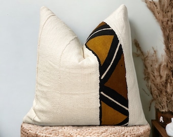 May include: A white throw pillow with a geometric pattern in brown, black, and yellow on one side. The pillow is sitting on a woven stool.