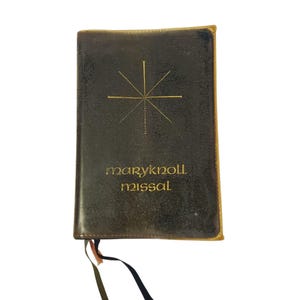 May include: A dark brown leather-bound book with gold lettering and a starburst design. The text "maryknoll missal" is printed in gold. A ribbon bookmark is visible.