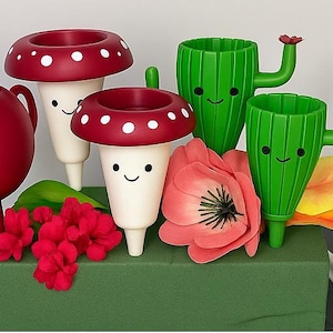 May include: A quirky display of a red teapot, mushroom-shaped planters with white spots, and green cactus-shaped planters, all with smiling faces. A large pink flower and red flowers are also included in the arrangement.