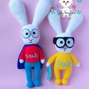 Könnte beinhalten: Zwei handgefertigte Häkelkaninchen-Puppen. Eines trägt einen roten Umhang, blaue Hosen und ein rotes Hemd mit dem Namen "Santi". Das andere trägt einen gelben Overall und eine schwarze Brille, ebenfalls mit dem Namen "Santi".