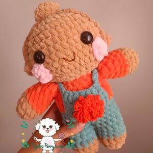 Pode incluir: Um brinquedo de pelúcia de crochê com pelo marrom, uma camisa vermelha e um macacão azul. O brinquedo tem um rosto sorridente com olhos pretos e bochechas rosadas.