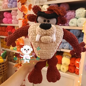 Może przedstawiać: Ręcznie robiona, pluszowa lalka Diabła Tasmańskiego w odcieniach brązu, beżu, czerni i bieli. Lalka na szydełku ma charakterystyczne cechy postaci, w tym duże zęby i kępkę włosów. Lalka jest trzymana przed półką z kolorową przędzą.