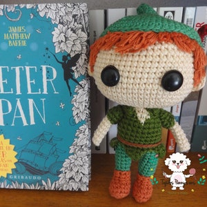 Puede incluir: Un muñeco de Peter Pan de crochet con un sombrero verde, cabello castaño y zapatos marrones. El muñeco está de pie frente a un libro titulado "Peter Pan" de James Matthew Barrie.