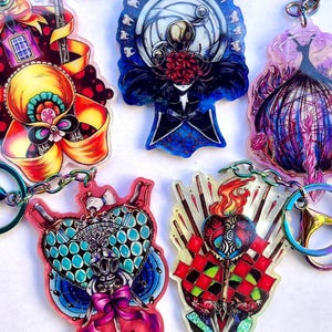 May include: A collection of five colorful acrylic keychains with intricate designs. Each keychain features a unique illustration, including a bow, a figure with a moon, and a heart. The keychains have iridescent hardware and are connected by silver chains.