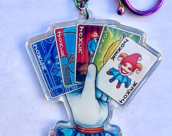 May include: A keychain featuring a hand holding playing cards with a joker theme. The cards display the word "Joker" and a cartoon jester. The keychain has a rainbow-colored ring and clasp.