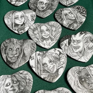 May include: A collection of heart-shaped pins featuring black and white ink illustrations of various fantasy characters, including humanoids, creatures with horns, and figures with multiple faces, all displayed in a grid-like arrangement.