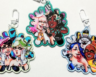 May include: Three colorful keychains featuring cartoon characters from the video game Splatoon. Each keychain is shaped like a splatter of ink and features two characters. The first keychain is teal and features a pink and white character with a microphone and a green and black character with headphones. The second keychain is light green and features a pink and black character with a microphone and a green character with a microphone. The third keychain is light blue and features a blue character with a microphone and a yellow character with headphones.