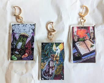May include: Three acrylic keychains with gold moon-shaped charms. Each keychain features a different image: a smudging stick with smoke, a glass bottle with a lit candle inside, and an open book with a "Good Luck Spell" inscription.