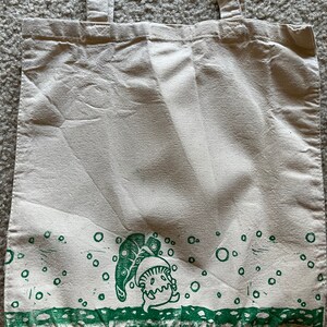 May include: A white canvas tote bag with green handles and a green print of a cartoon character with a leaf on its head. The print is surrounded by green circles.