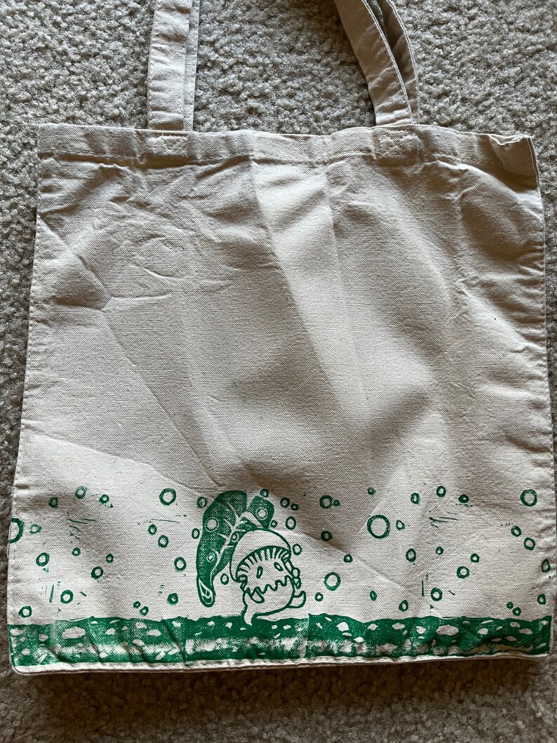 May include: A white canvas tote bag with green handles and a green print of a cartoon character with a leaf on its head. The print is surrounded by green circles.