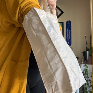 May include: A person is holding a white canvas tote bag with two handles. The bag is folded over at the top and has a slight crease down the middle.