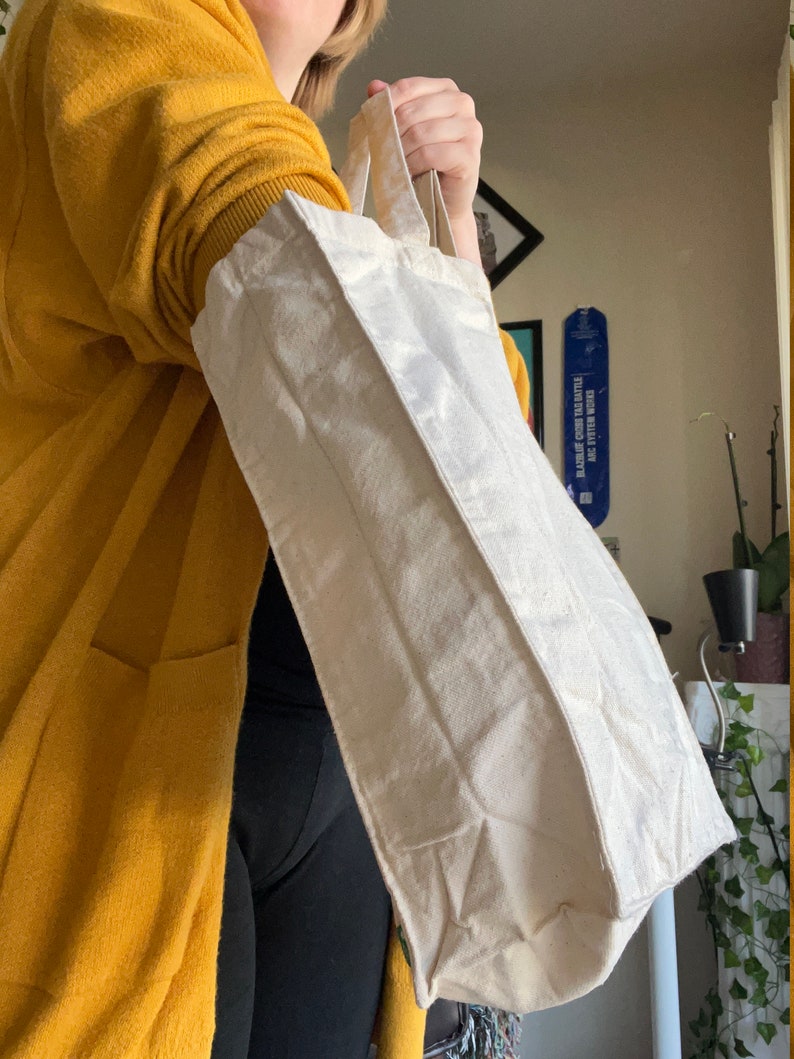 May include: A person is holding a white canvas tote bag with two handles. The bag is folded over at the top and has a slight crease down the middle.