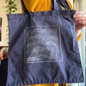May include: A dark blue tote bag with a white graphic of a woman's face and waves. The graphic is in a square frame.