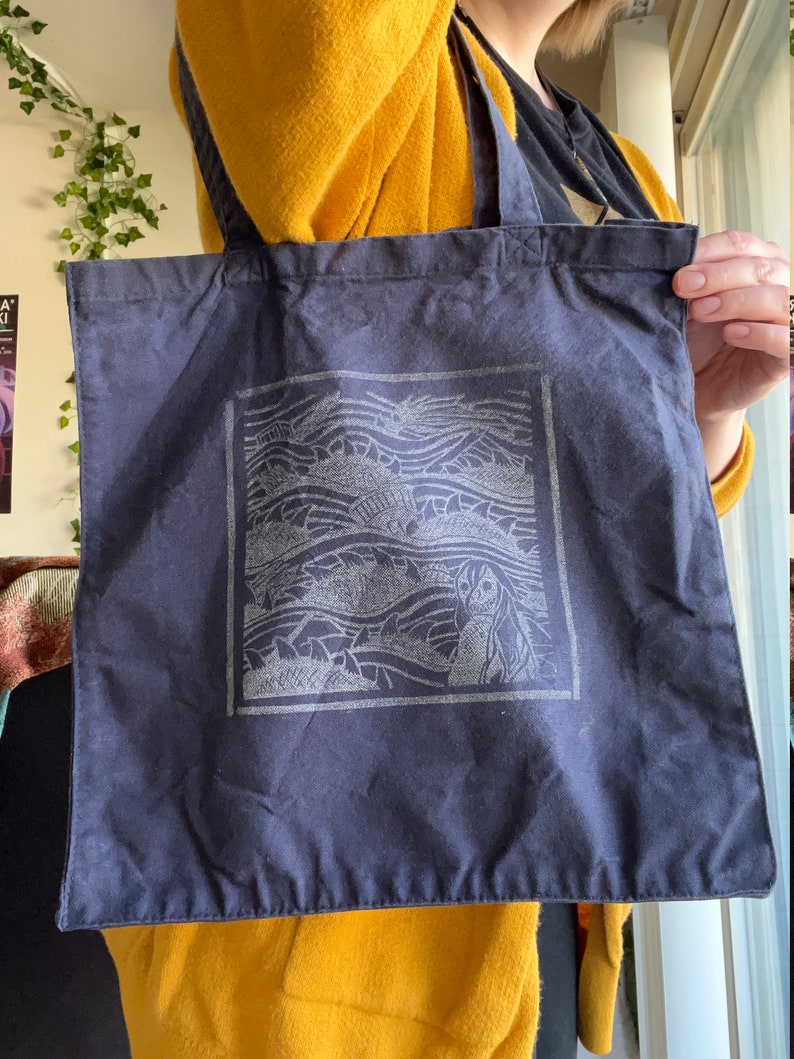 May include: A dark blue tote bag with a white graphic of a woman's face and waves. The graphic is in a square frame.