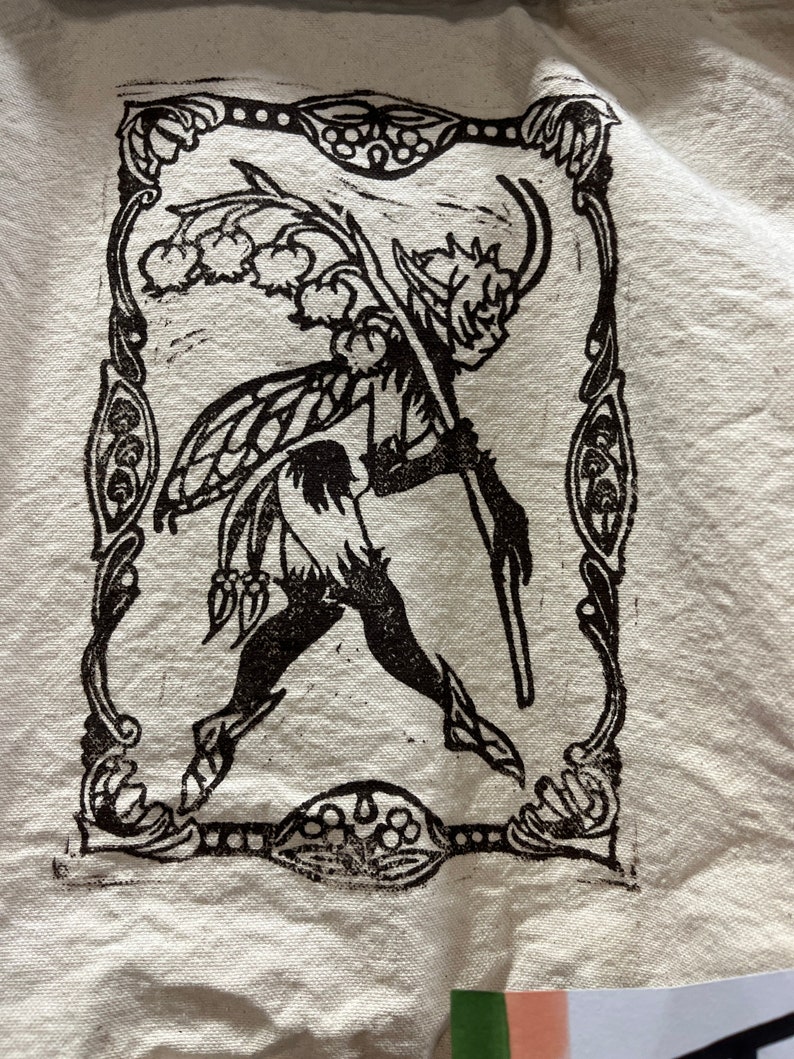 May include: Black and white illustration of a fairy-like creature with wings, holding a staff with flowers. The illustration is framed with a decorative border.