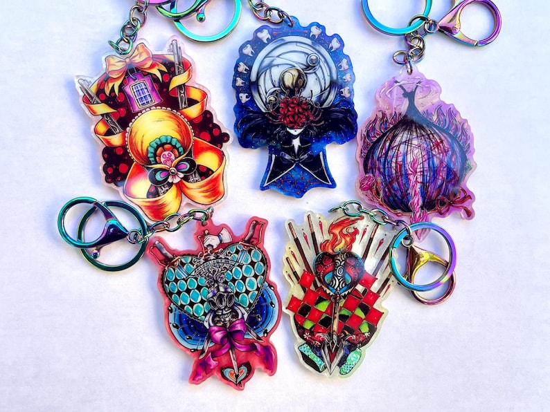 May include: A collection of five colorful acrylic keychains with intricate designs. Each keychain features a unique illustration, including a bow, a figure with a moon, and a heart. The keychains have iridescent hardware and are connected by silver chains.