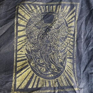 May include: Black fabric tote bag featuring a gold-coloured graphic print of a stylised winged creature with eyes. The graphic is surrounded by a sunburst design.